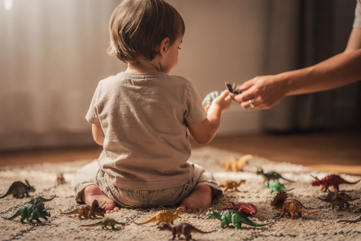 Kleinkind von hinten sitzt auf einem Teppich mit Dinosauriern, eine erwachsene Hand reicht eine Figur herüber