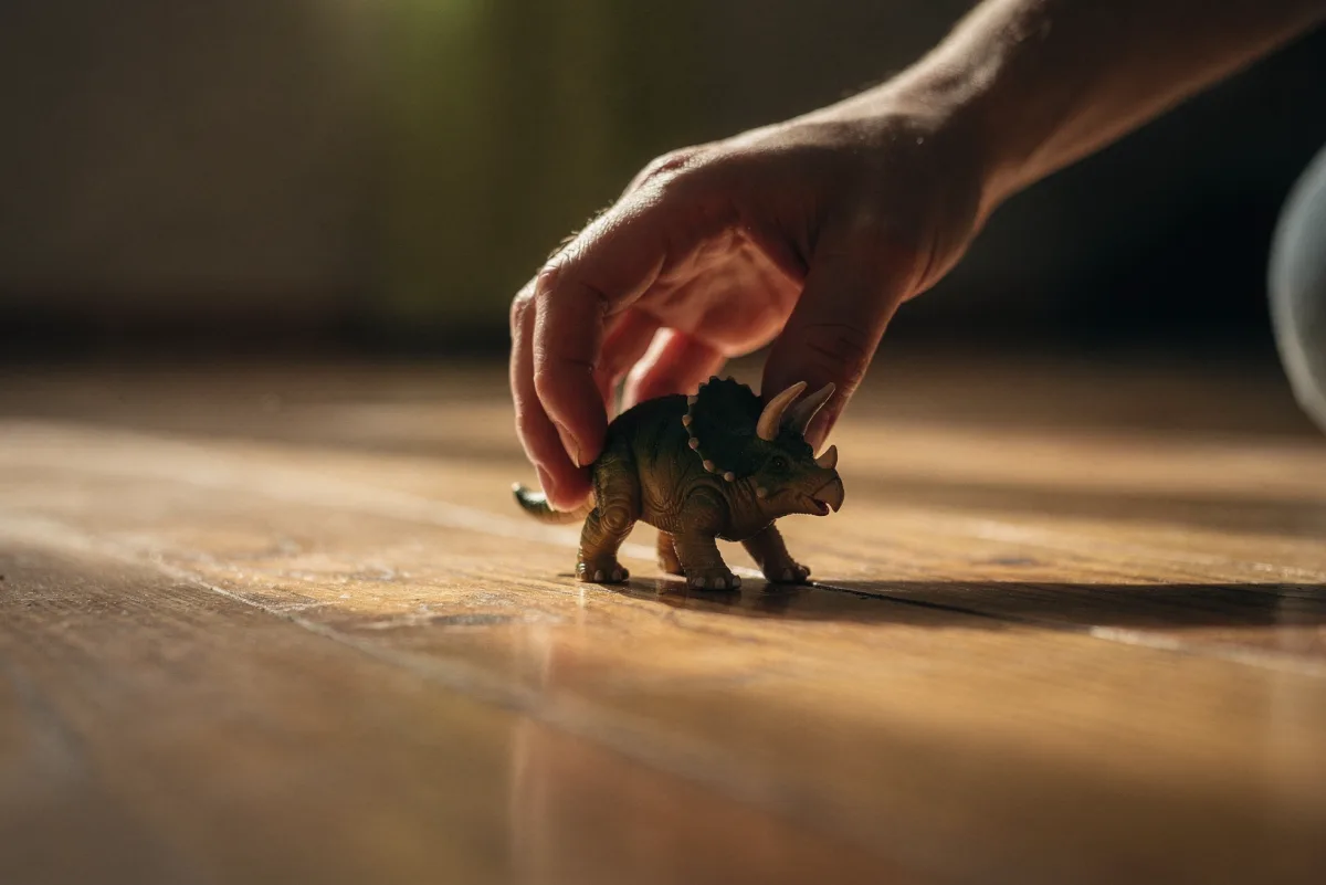 Hand nimmt eine kleine Triceratops-Figur vom Holzboden auf, warmes Licht und weicher Hintergrund