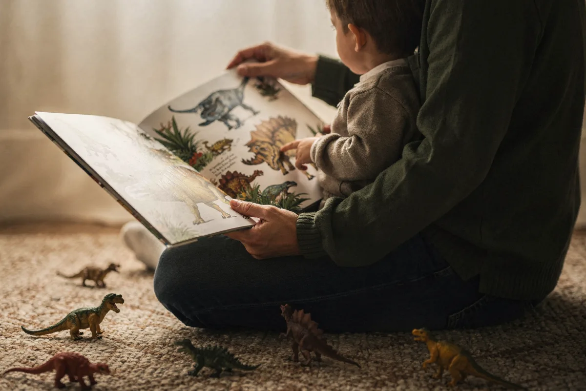 Elternteil und kleines Kind sitzen zusammen auf einem Teppich und schauen ein illustriertes Dinosaurierbuch an, Plastikdinos liegen um sie herum