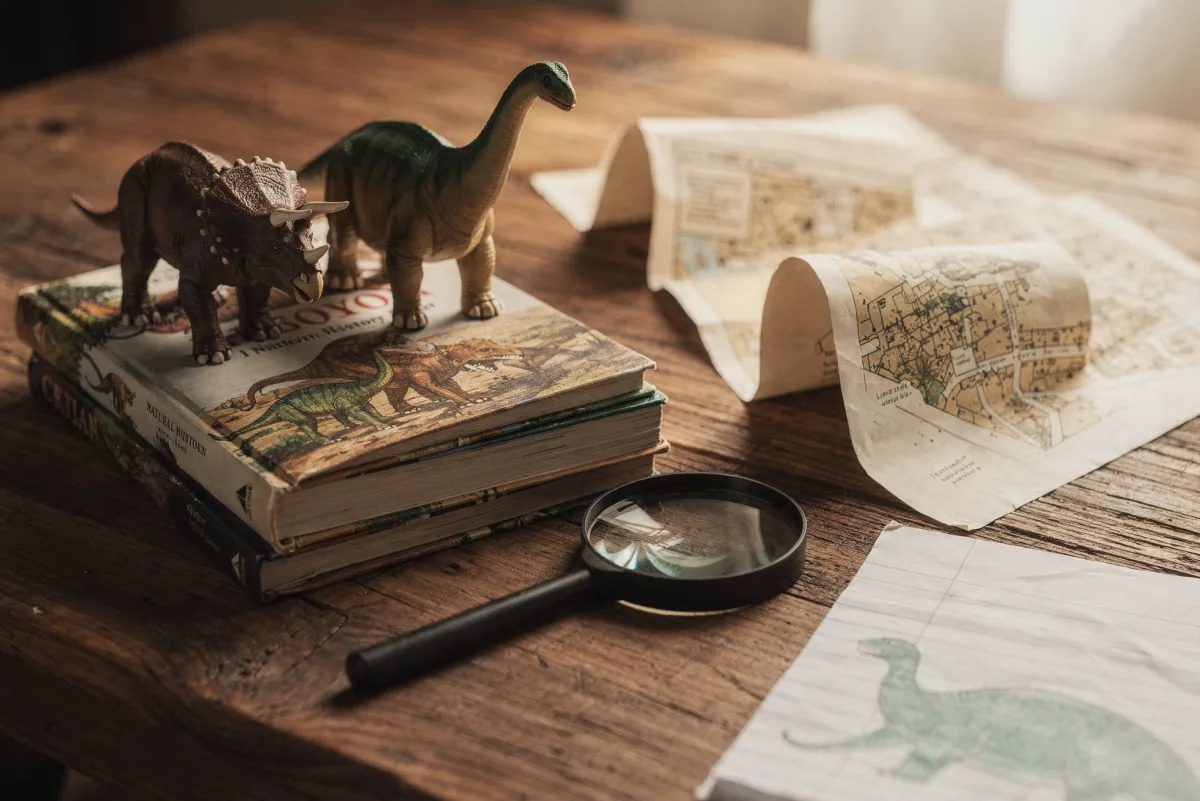 Triceratops- und Brachiosaurus-Figuren auf einem Stapel Dinosaurier-Sachbücher neben einer aufgerollten Karte, einer Lupe und einer Kinderzeichnung