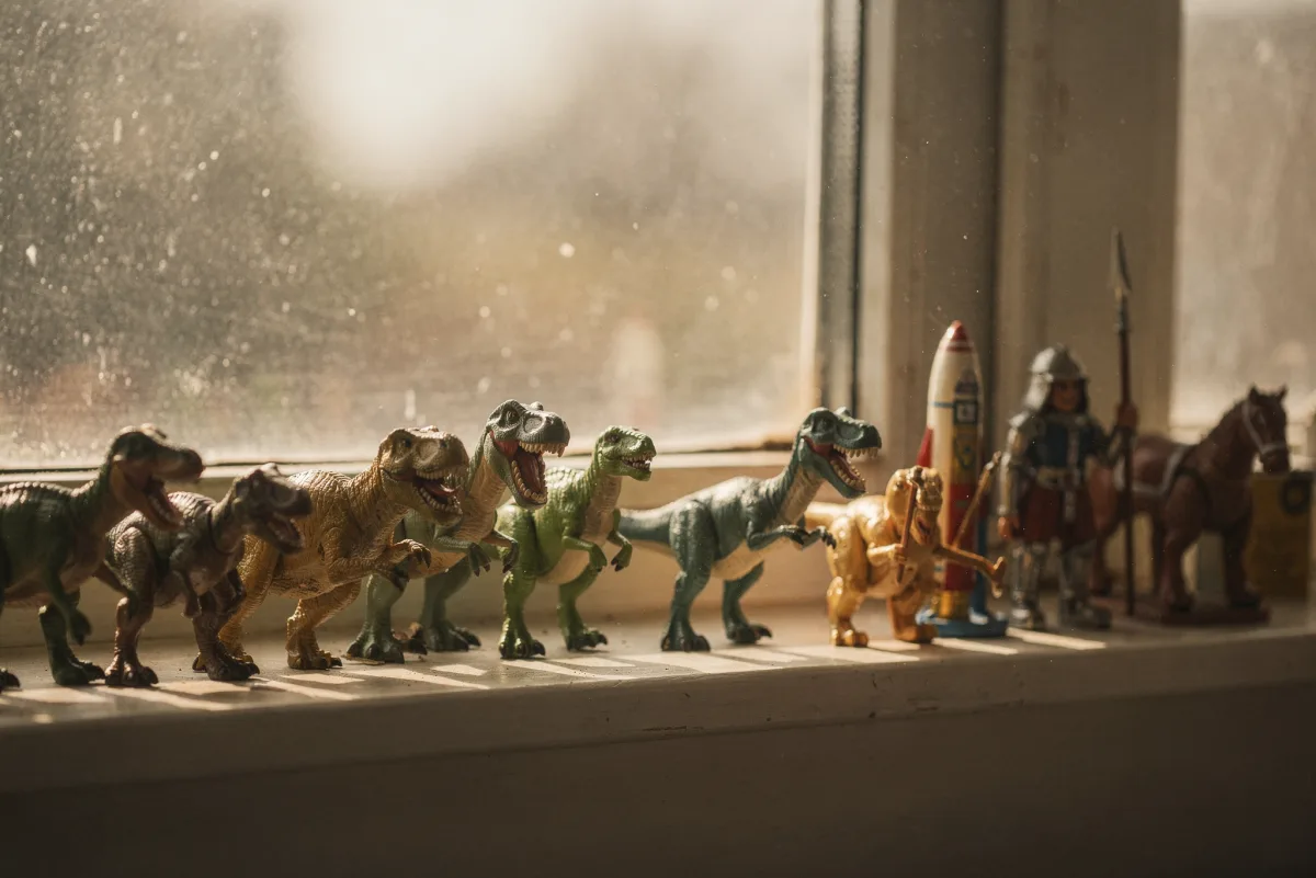Eine Reihe Spielzeug-Dinosaurier auf einem staubigen Fensterbrett, gefolgt von einer Rakete, einem Ritter und einem Pferd im warmen Gegenlicht
